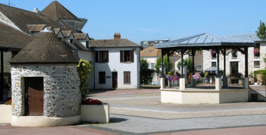 Place de village ensoleillée avec kiosque fleuri, bâtiments traditionnels et petite tour en pierre
